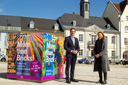 Caption: Museum director Anna Dziwetzki and 1st mayor Michael Abraham of the city of Rehau with the advertising cube of the special exhibition "More than Bricks!" on the Rehauer Maxplatz.